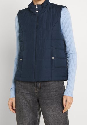 Personne portant une veste matelassée bleu marine avec fermeture éclair et poches à pression, par-dessus un haut à manches longues côtelé bleu clair et un jean gris.