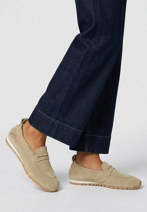 Beige suède instap loafers gedragen met donkerblauwe jeans met rechte pijpen, met zichtbare zijkant- en zooldetails.