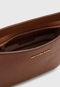 Bruine Michael Kors-handtas met een gladde textuur, ritssluiting in goudkleur en geperst logo aan de voorkant. Binnenvak zichtbaar.