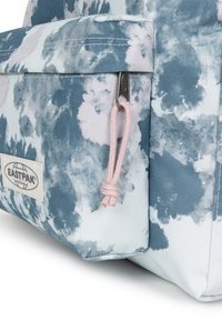 Mochila azul y blanca con un patrón de tie-dye, que cuenta con un bolsillo con cremallera y un lazo de cuerda rosa. El material parece duradero y con textura.