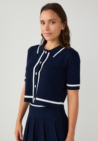 Marinefarbener Kurzarm-Cardigan mit Kragen, zusätzlich mit weißen Abschlüssen und Knöpfen. Strukturierter Stoff, cropped Design und taillierte Passform.