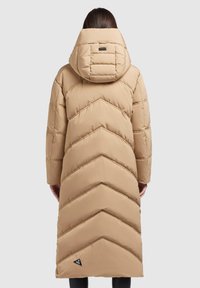 Manteau d'hiver long beige matelassé avec une grande capuche, motif de couture en chevrons, porté par une personne se tenant de dos.