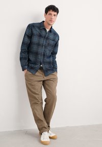 Man leunt tegen een witte muur, gekleed in een blauw geruiten overhemd, beige broek en witte sneakers, met zijn handen in zijn zakken, kijkend recht vooruit.