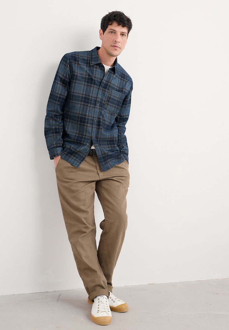 Man leunt tegen een witte muur, gekleed in een blauw geruiten overhemd, beige broek en witte sneakers, met zijn handen in zijn zakken, kijkend recht vooruit.