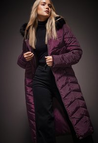 Lang, polstret frakke i dyb lilla med en kunstig pels hætte, med diagonalt kviltet design og sidelommer med lynlås. Båret over en sort bluse og bukser.