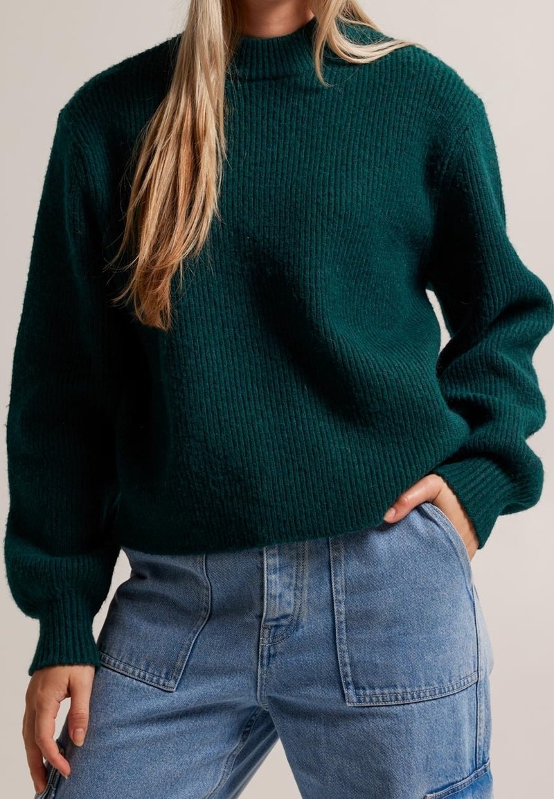 Pull en tricot côtelé vert avec un col montant, présentant une coupe ample et des épaules tombantes, associé à un jean taille haute.