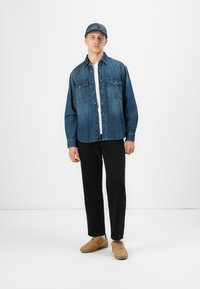Jeune homme portant une chemise en denim bleu et une casquette, un pantalon noir et des baskets beiges, se tenant devant un fond blanc uni.