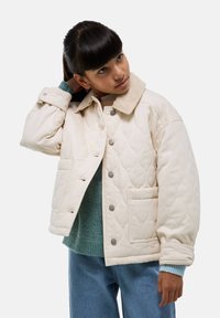 Veste beige matelassée avec un col en velours côtelé, boutons-pression et deux poches avant. Portée par-dessus un pull vert et un jean bleu.