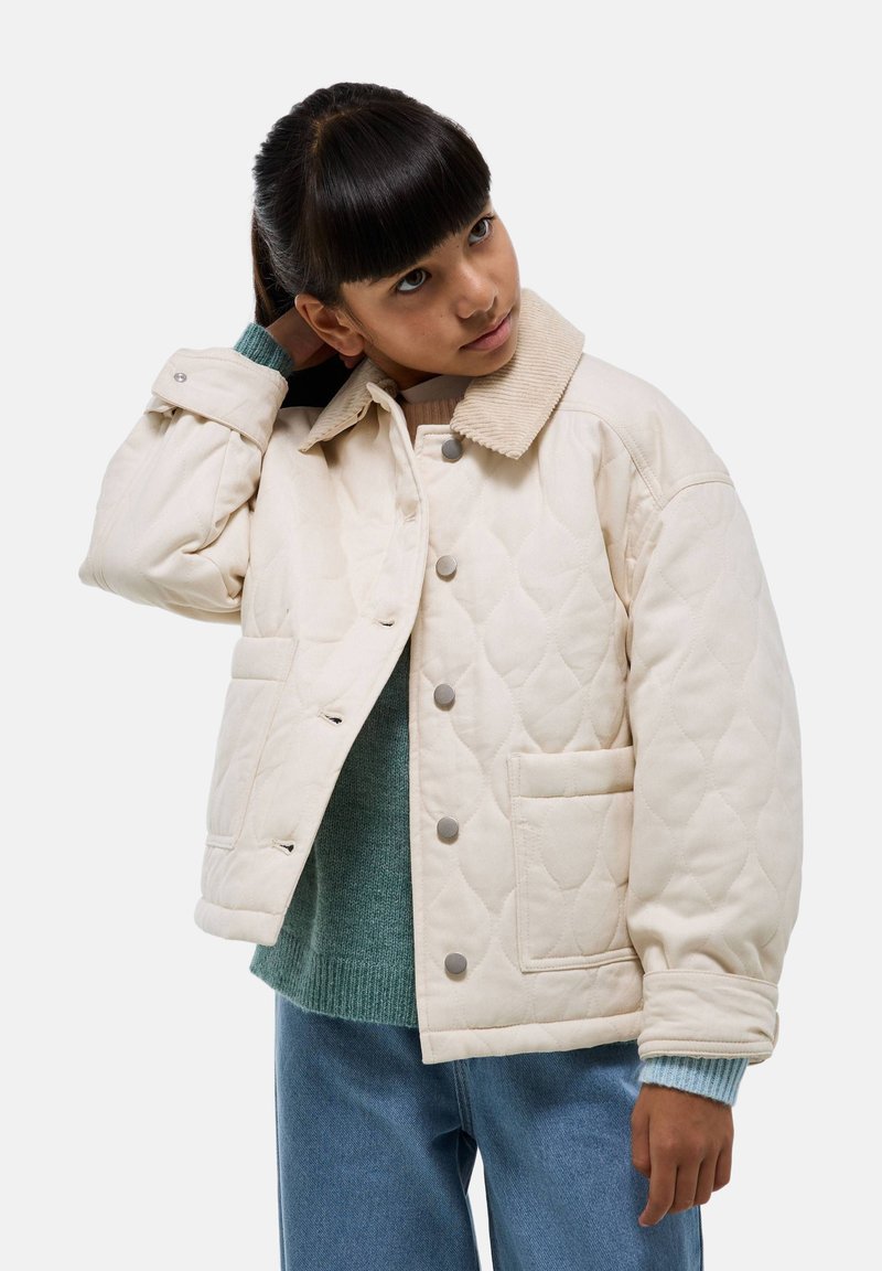 Veste beige matelassée avec un col en velours côtelé, boutons-pression et deux poches avant. Portée par-dessus un pull vert et un jean bleu.