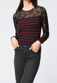 Bordeaux-rode en zwarte gestreepte top met lange zwarte kanten mouwen, ronde halslijn en ribbelmanchetten. Draag het met donkere jeans, handen in de zakken.