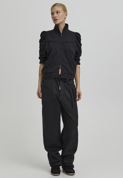 Žena u crnoj jakni s punim rukavima na patentni zatvarač i širokim crnim hlačama stoji i gleda u stranu na jednobojnoj sivoj pozadini.