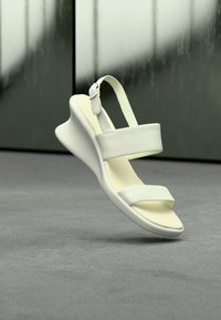 Λευκό πέδιλο ανοιχτού δακτύλου με χοντρές λουρίδες και αγκράφα, με ανυψωμένη, καμπύλη σόλα πάνω σε θολό πράσινο και γκρι φόντο.