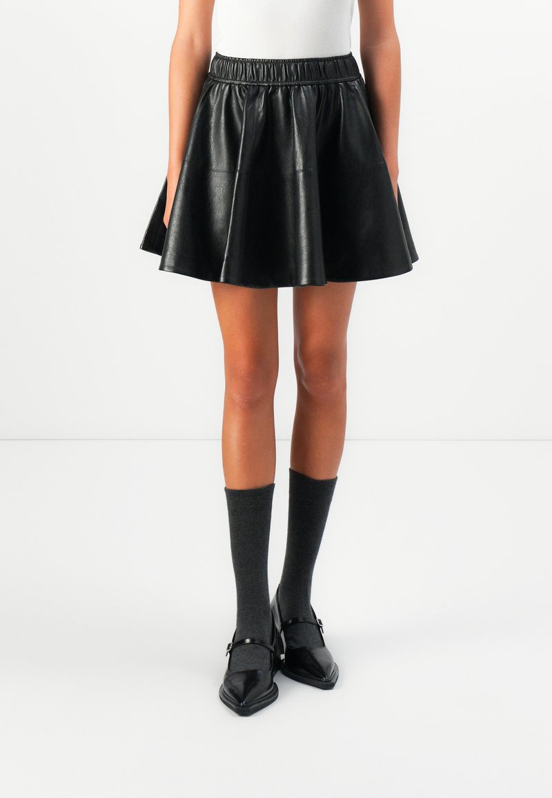 Person wearing a black leather flared mini skirt, dark gray knee-high socks, and black Mary Jane heels standing on a white floor.