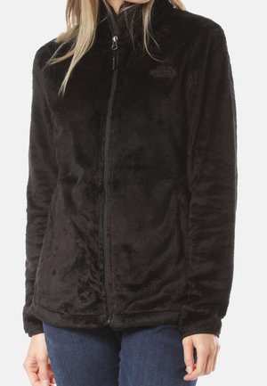 Vrouw draagt een zwarte fleecevest met rits, handen ontspannen langs haar zij, gecombineerd met een blauwe spijkerbroek.