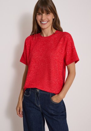 Lachende vrouw die een rood truitje met korte mouwen draagt, met één hand in de zak van een donkerblauwe spijkerbroek tegen een effen achtergrond.