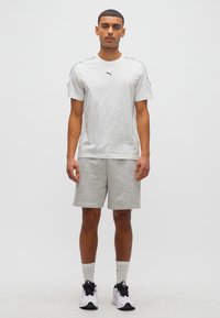 Άνδρας που στέκεται φορώντας άσπρο t-shirt Puma, γκρι σορτς, άσπρες κάλτσες και άσπρα αθλητικά παπούτσια, σε απλό λευκό φόντο.