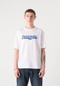 White cotton T-shirt with a large, colorful "hug" graphic in blue and black. Casual fit, short sleeves, round neck, and denim jeans.