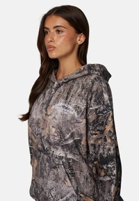 Hoodie à motif camouflage en tissu doux. Doté d'une grande poche et d'une capuche avec cordon de serrage. Les couleurs incluent des nuances de marron, noir et vert.