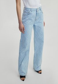 Jean taille haute bleu clair avec un motif en zigzag, coupe droite et coutures contrastantes. Porté avec des sandales à talons noires.