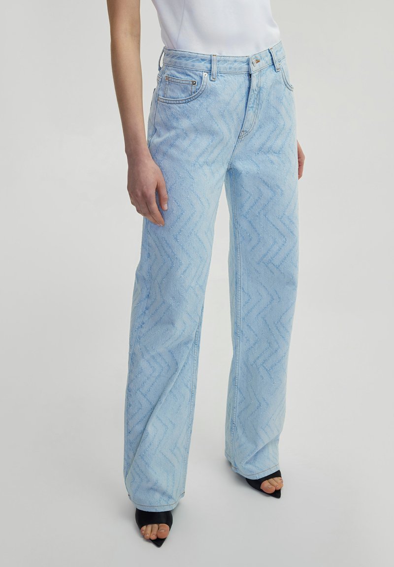 Jean taille haute bleu clair avec un motif en zigzag, coupe droite et coutures contrastantes. Porté avec des sandales à talons noires.