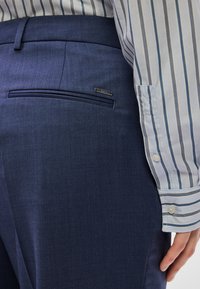 Στενά μπλε σκούρα παντελόνια με υφή, με μία τσέπη στο πίσω μέρος και μια μικρή διακοσμητική μεταλλική ετικέτα. Φαίνεται η ριγέ μανσέτα του πουκάμισου.