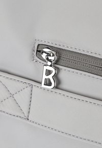 Coin de sac en tissu gris clair avec une poche zippée. Caractéristiques en métal argenté avec un tirette en forme de "B". Des accents incluent des coutures texturées.