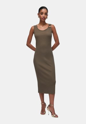 Donna che indossa un abito midi senza maniche verde oliva con dettaglio ad anello sulla spalla sinistra e sandali dorati con tacco alto, in piedi con le mani dietro la schiena.