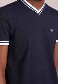 Námořnická modrá polo tričko s V-výstřihem a bílými lemy. Krátké rukávy mají bílé pruhy. Malý loga na hrudi. Bavlněný materiál.