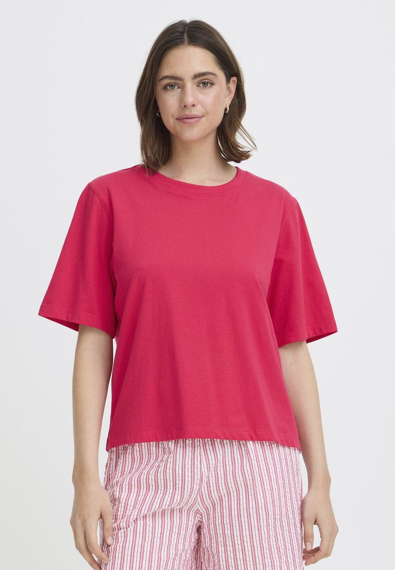 Femme aux cheveux bruns mi-longs portant une chemise rouge ample à manches courtes et un pantalon blanc à rayures verticales rouges, debout devant un fond blanc.