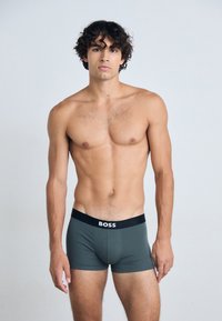 Dunkelgrüne Baumwoll-Boxershorts mit einem schwarzen elastischen Bund, der ein weißes Logo trägt. Das Modell wird mit freiem Oberkörper und neutralem Hintergrund gezeigt.