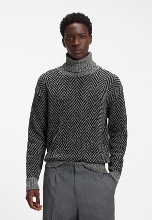 Mand iført en sort og hvid turtleneck sweater med chevronmønster og grå teksturerede bukser, stående med den ene hånd i lommen mod en ensfarvet baggrund.