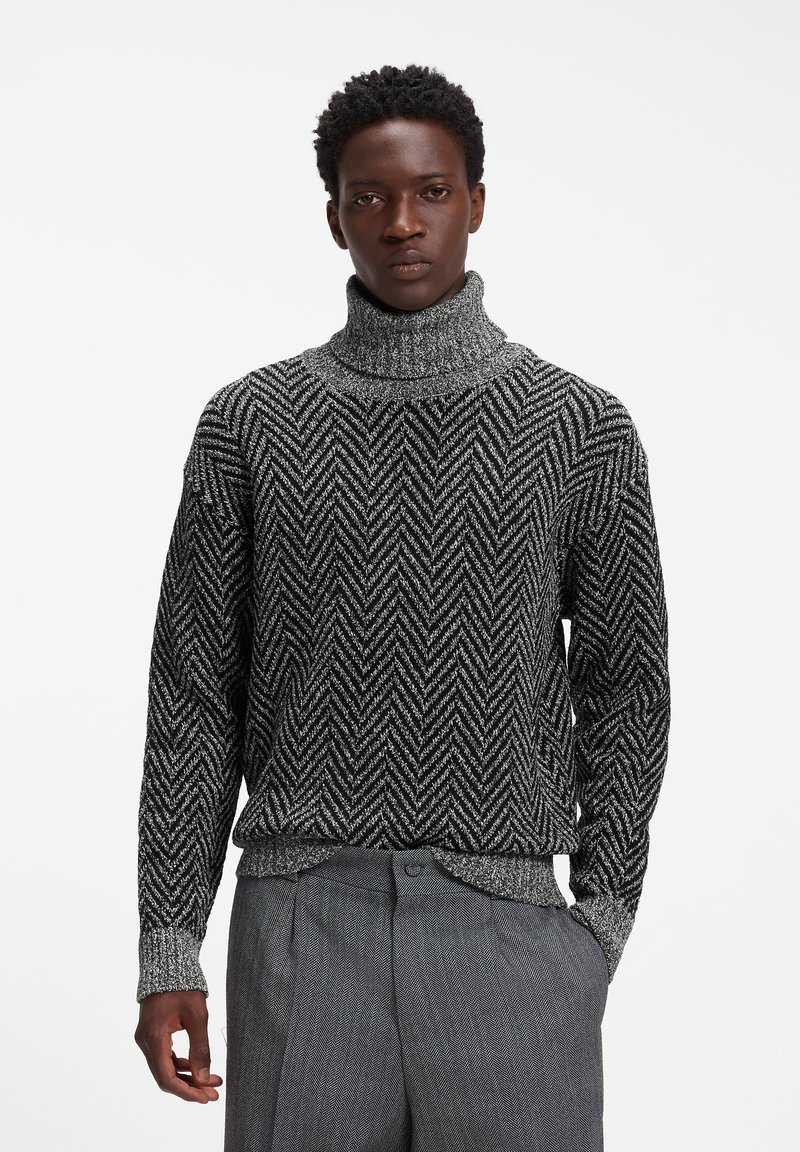 Homme portant un pull à col roulé à motif chevrons noir et blanc et un pantalon gris texturé, debout avec une main dans la poche devant un fond uni.