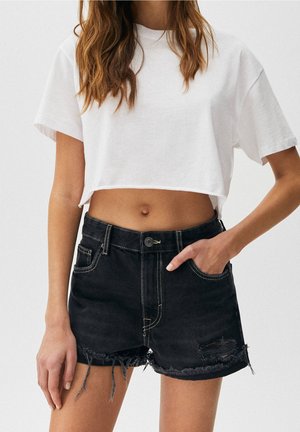 Femme portant un t-shirt court blanc et un short en jean noir déchiré, debout, une main dans la poche, sur un fond uni.