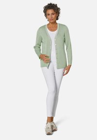 Hellgrüne gerippte Strickjacke mit V-Ausschnitt und Knopfverschluss, kombiniert mit einem weißen Oberteil und schmalen weißen Hosen, getragen mit beigen Sneakers.