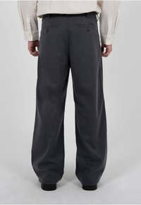 Homme portant un pantalon gris foncé ample et une chemise rayée claire rentrée, se tenant sur un fond uni gris clair.