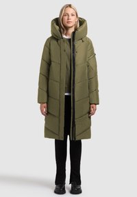 Abrigo acolchado verde oliva con una gran capucha, cierres de cremallera y botón a presión, patrón de rombos acolchado y bolsillos laterales, usado sobre pantalones negros y botas.