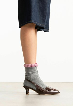 Been met een donkerbruine schoen met middelhoge hak, gecombineerd met een grijze geribbelde sok met roze ruches, en een donker denim cropped broek.