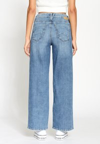 Žena ve světlemodrých širokých džínách a bílých teniskách, stojící zády k jednoduchému pozadí.