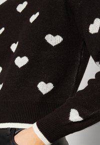 Črn pleten pulover z belimi srčastimi vzorci. Rebrasti manšeti in rob z kontrastnim belim obrezom. Mehka tekstura in sproščen kroj.