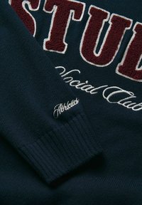 Jūras spēku džemperis ar sarkaniem un baltajiem izšūtajiem vārdiem "STUD" un "Athletics". Mīksta tekstūra, ribotas manšetes, slīpa skata leņķis, kas izceļ detaļas.