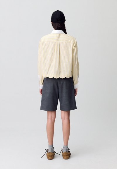 Camicia gialla a righe con orlo scallop, abbinata a pantaloni grigi. Modello che indossa calzini grigi e scarpe marroni, visto da dietro.