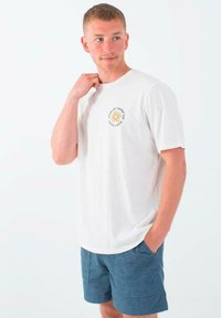 Camiseta de algodón blanca con escote redondo y un pequeño gráfico en el frente de un sol con texto. Combinada con pantalones cortos azules texturizados.