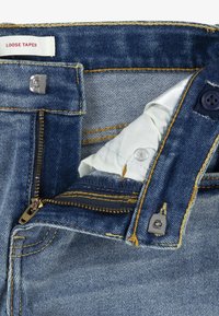 Lösa tapered denim jeans med dragkedja och knappstängning, kontrasterande guldtråd och en ljusblå tyginredning med varumärkesetikett.