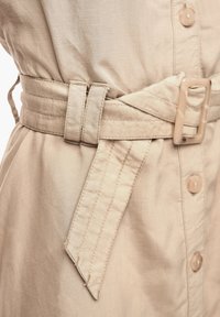 Beige klänning i tyg med midja som betonas av ett bälte med rektangulär spänne, knappdetaljer och en strukturerad yta.
