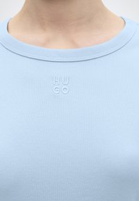 Κοντινή λήψη του λαιμού και του άνω μέρους του στήθους με ανοιχτό μπλε πουκάμισο με στρογγυλή λαιμόκοψη και μικρό ανάγλυφο λογότυπο "HUGO" στο κέντρο κάτω από το γιακά.