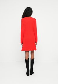 Femme aux cheveux bouclés portant une robe rouge vif à manches longues côtelée et des bottes noires montant jusqu'aux genoux, de dos sur un fond blanc uni.