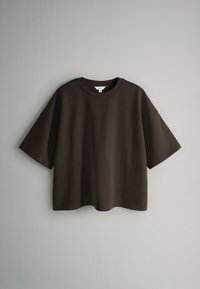 Tricou oversized maro închis cu mâneci scurte şi guler rotund, atârnat pe un fundal simplu gri deschis.