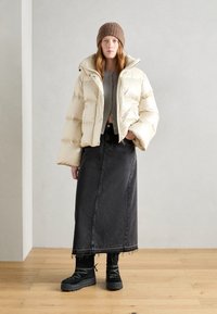 Veste crème duveteuse, crop top gris, jupe en denim noire longue, et bottes noires imposantes avec des détails texturés ; bonnet en tricot.