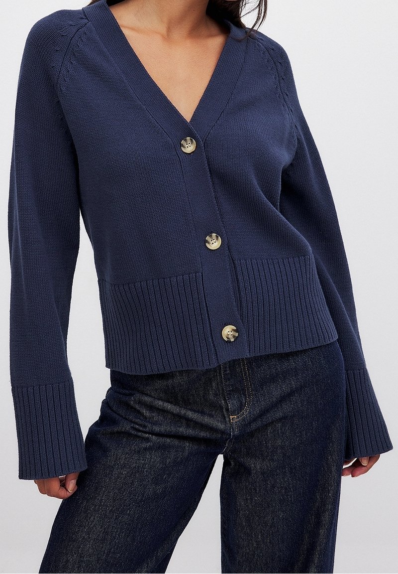 Granatowy kardigan z wycięciem w serek, ściągaczem u dołu i długimi rękawami. Posiada cztery złote guziki oraz ozdobne przeszycia na ramionach.