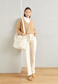 Beige strikket genser over en hvit krage skjorte, kremfargede bukser, brune joggesko og en lerrets skulderveske mot en nøytral bakgrunn.